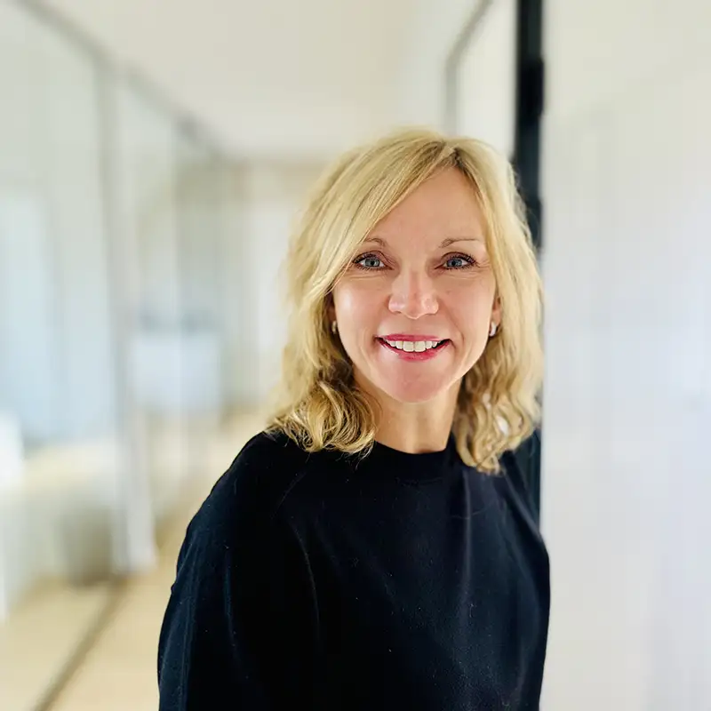 Une personne souriante avec des cheveux blonds porte un pull noir dans un environnement lumineux et moderne.